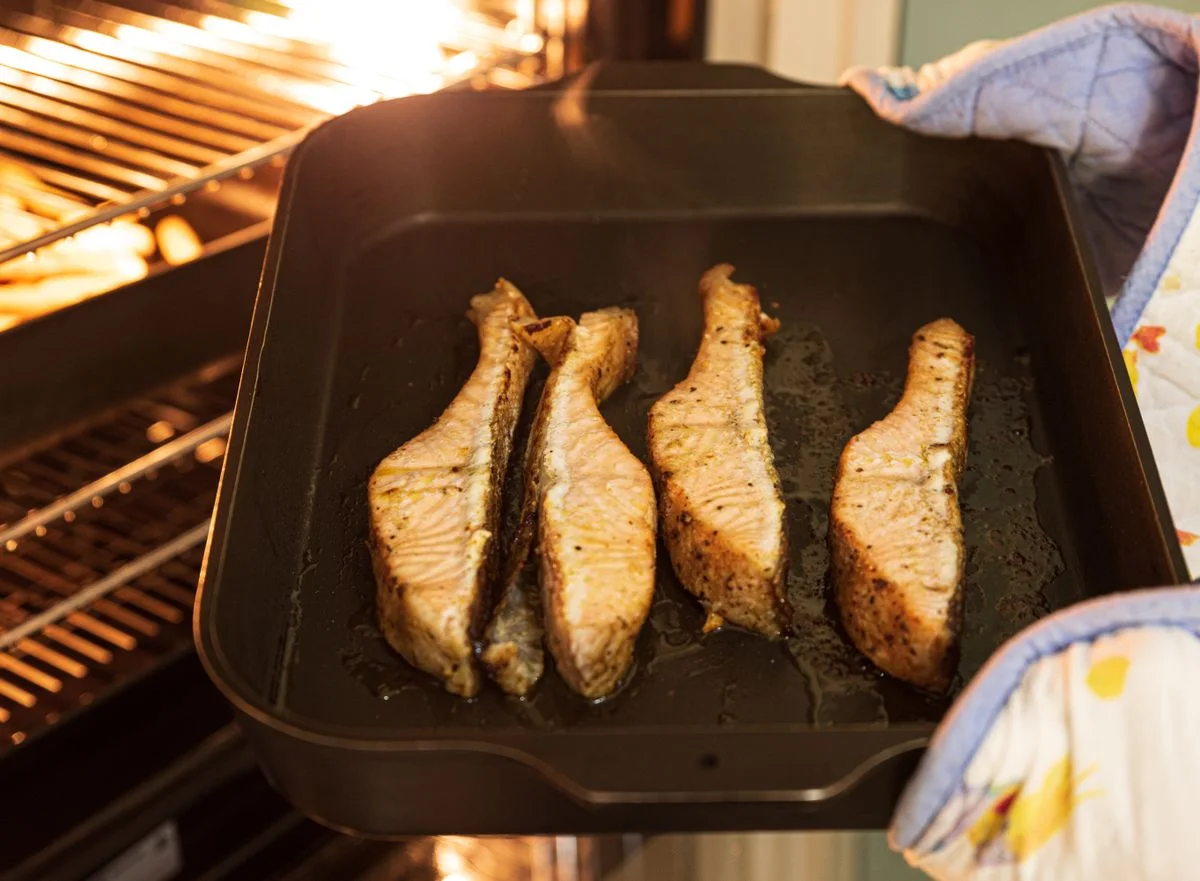 Close-up of four seasoned salmon fillets being baked in an oven with mitts.