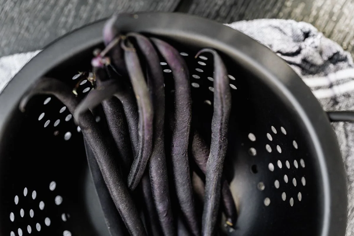 Fresh purple string beans in a black colander, ready for cooking.