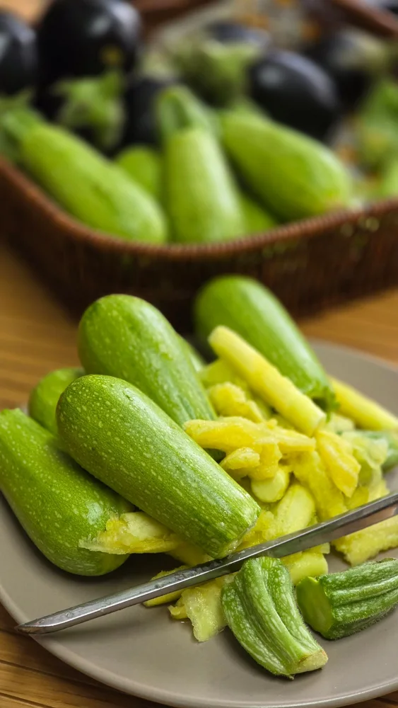 Close-up of fresh green zucchini and peeler on plate with basket of vegetables.