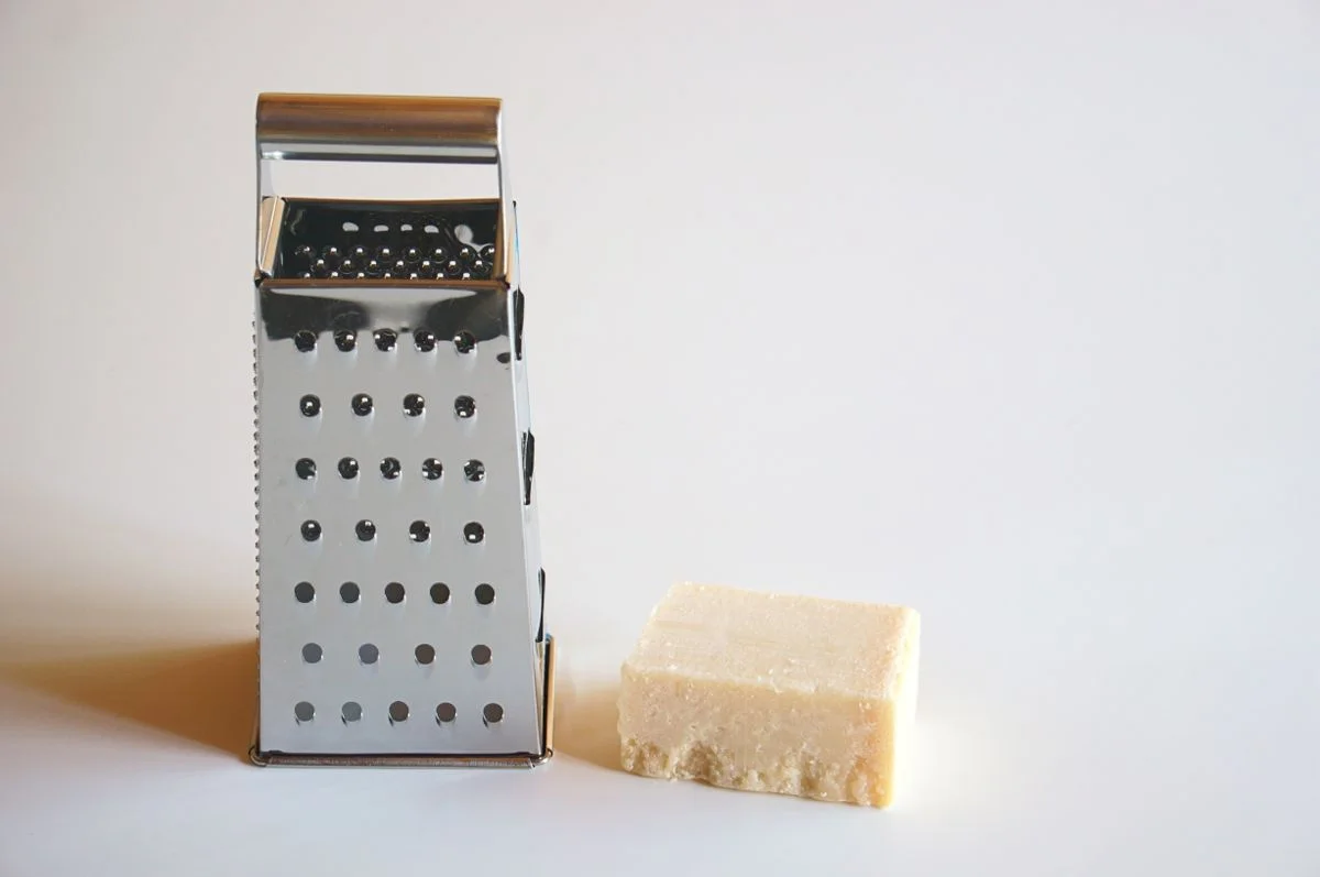 A stainless steel grater next to a block of parmesan cheese on a white surface.
