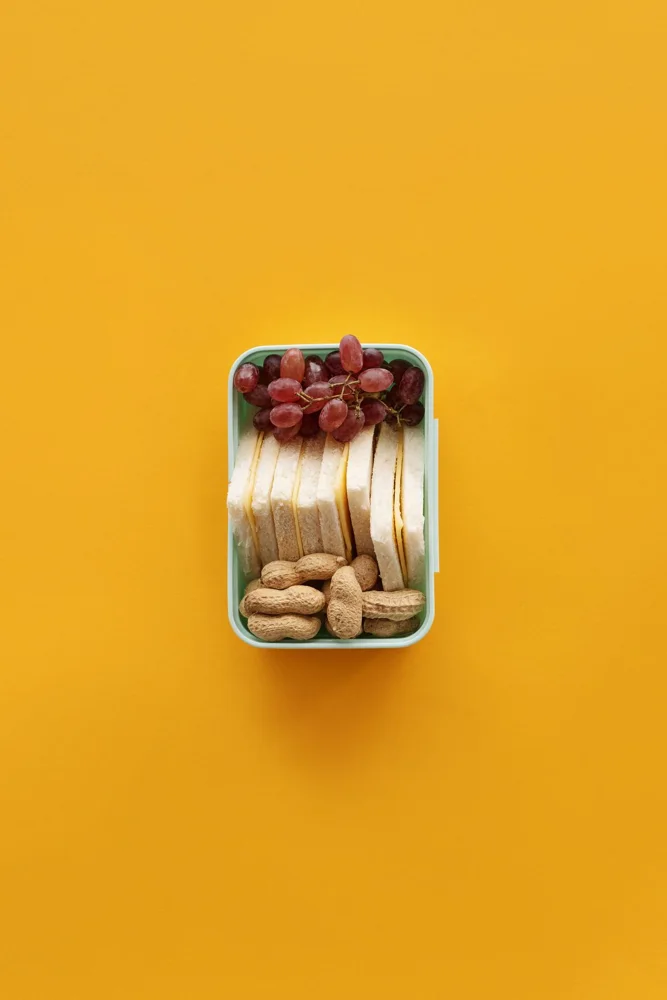 Top view of a lunch box with grapes, sandwich, and peanuts on yellow background.