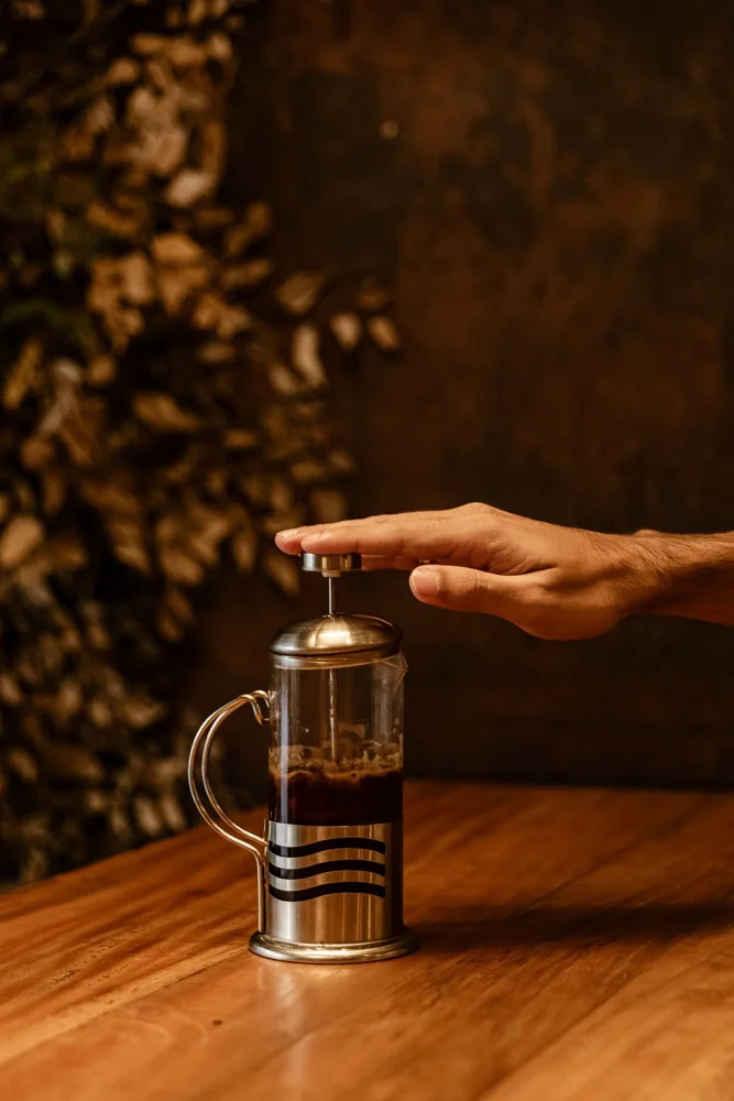 Hand pushing down on a French press coffee maker on a wooden table, warm and cozy atmosphere.