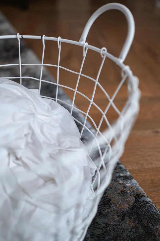 A white wire laundry basket with crumpled clothes placed on a hardwood floor inside a home.