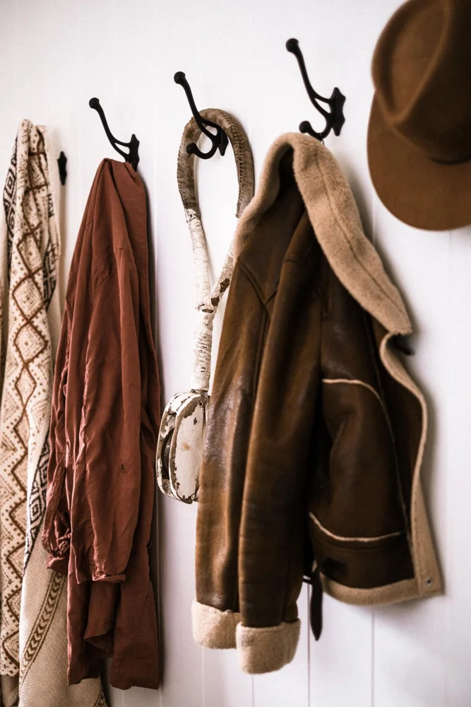 A warm leather coat and hat hang on stylish rustic hooks against a wooden wall.