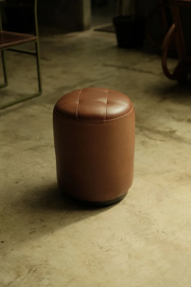 A cylindrical brown leather ottoman on a concrete floor in an indoor setting, ideal for modern interiors.