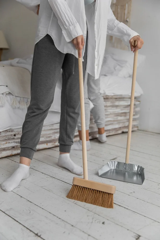 Crop unrecognizable female friends in comfy clothes sweeping floor using broom and dustpan while cleaning apartment during weekend