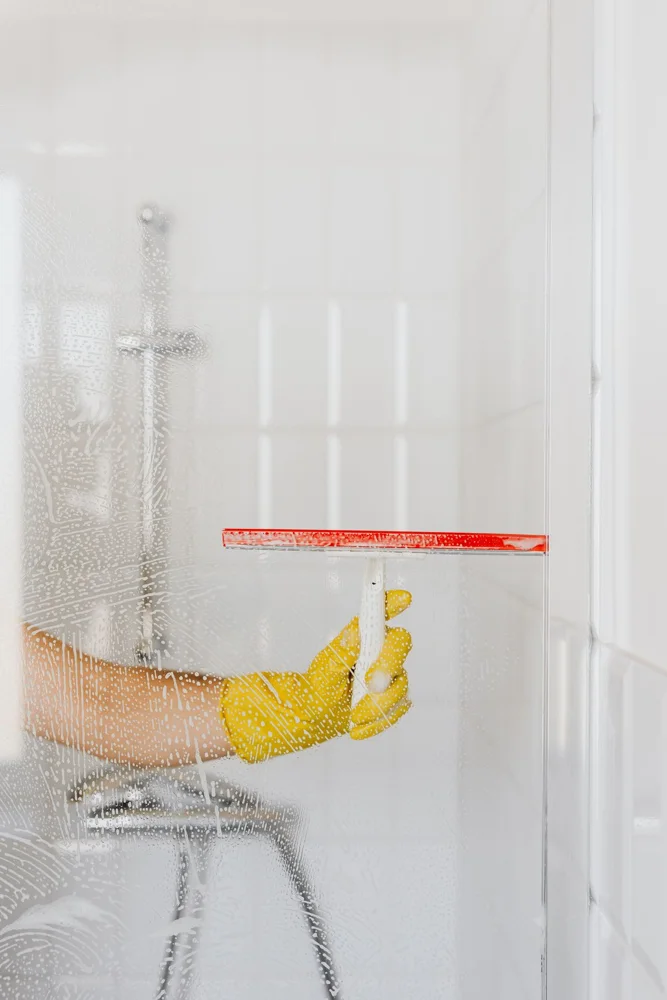 Crop anonymous person in yellow rubber glove cleaning glass shower unit with glass wiper and detergent against white tile wall