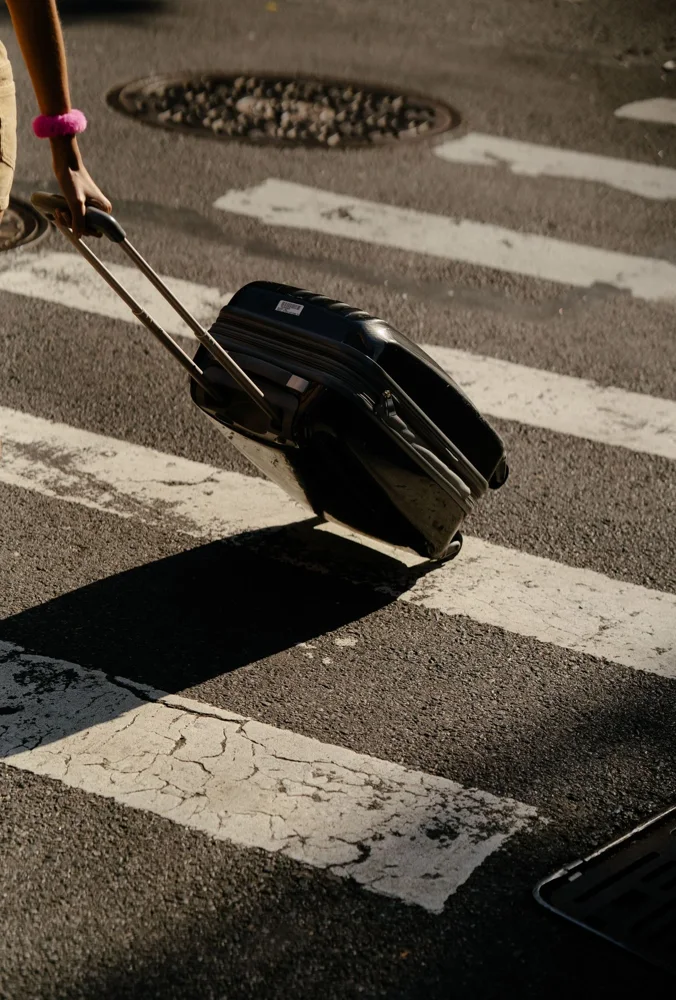 Person pulling suitcase across city crosswalk on a sunny day.