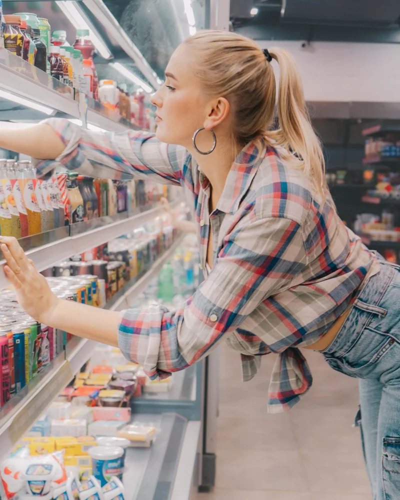Young woman reaches for a drink on a supermarket shelf. Casual and stylish shopping scene.