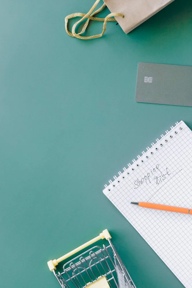 A shopping list on a spiral notebook with a bank card and mini cart on green background.