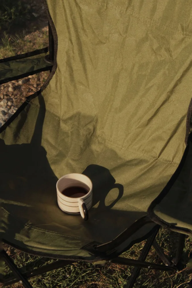 A cozy green camping chair holds a striped mug of coffee, basking in the outdoor morning light.